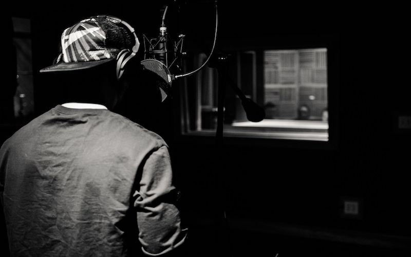 A black and white image of the back of a person wearing a backwards hat in front of a mic. The setting is inside a music studio.