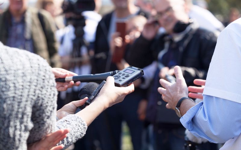 The forearms of reporters holding recording devices in front of a blurred out crowd.