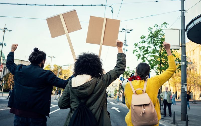 The image shows the backs of a group of protesters, walking down a street, holding signs and raising their fists.