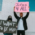 An image of two protesters holding up signs. The man on the right holds a sign that says "Justice 4 All". The woman on the left weras a face mask and her sign says "I want to be heard".